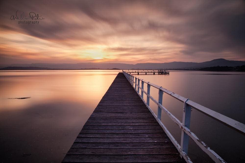 Sunset over lake illawarra with a pier leading into the sun.