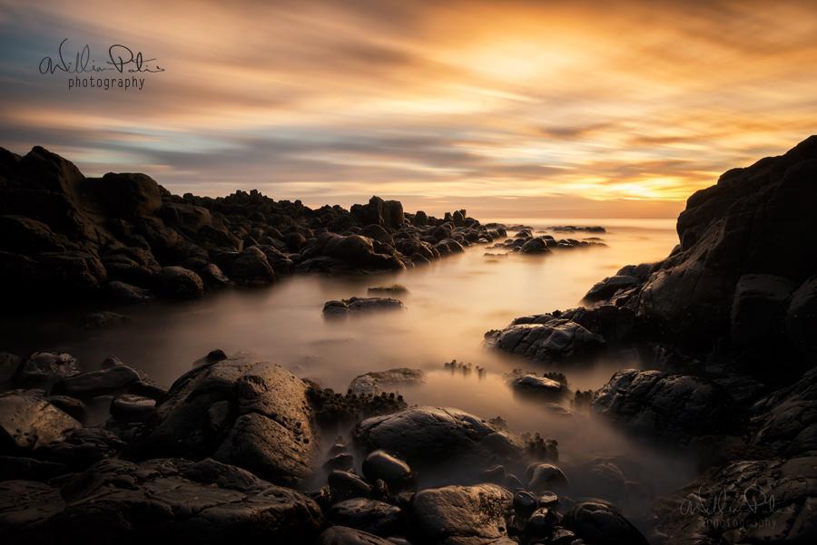 Rugged rocks upon the shore line are illuminated by the rising sun, surrounded by misty water.